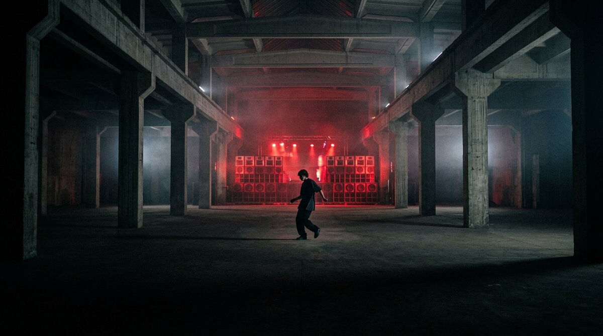 A single isolated dancer standing in darkness inside an industrial warehouse rave, distant red strobes and a stacked sound system creating tension